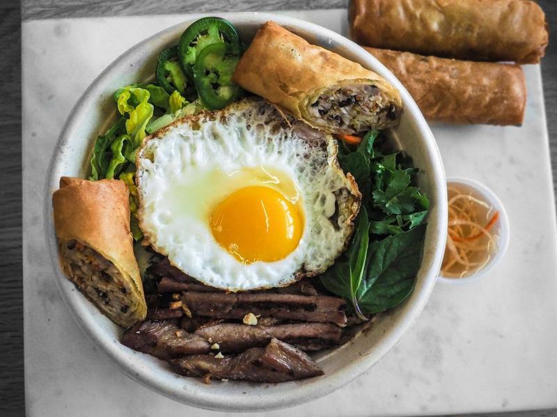 Fusion-style Vietnamese rice bowl with grilled meat, herbs, egg, and fried spring rolls